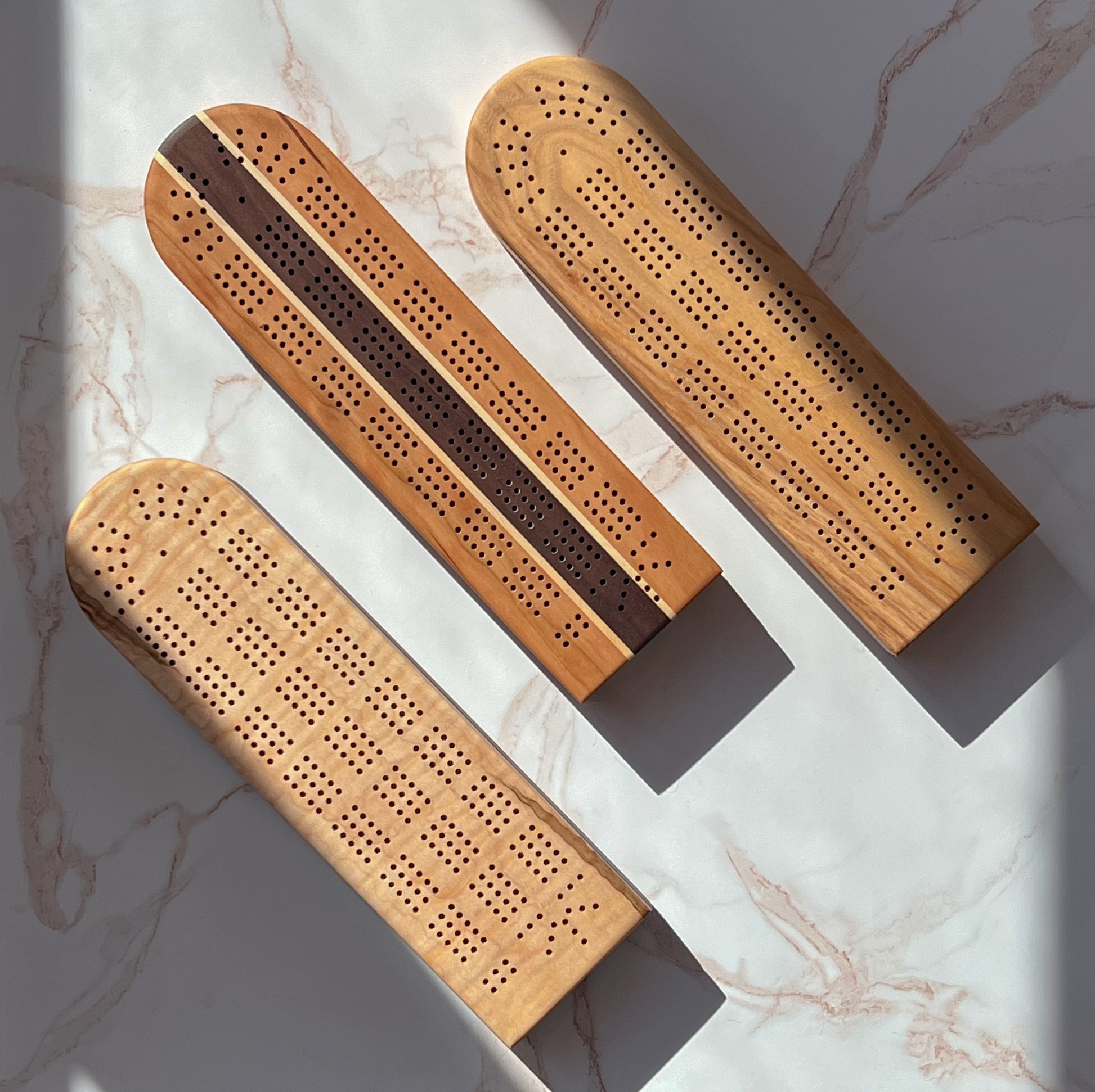 Three handmade wooden cribbage boards with swivel top card and peg storage in 3 different shades of natural wood, one has a dark wood stripe down the middle for an elegant twist made by Camino Woodshop in Milwaukee